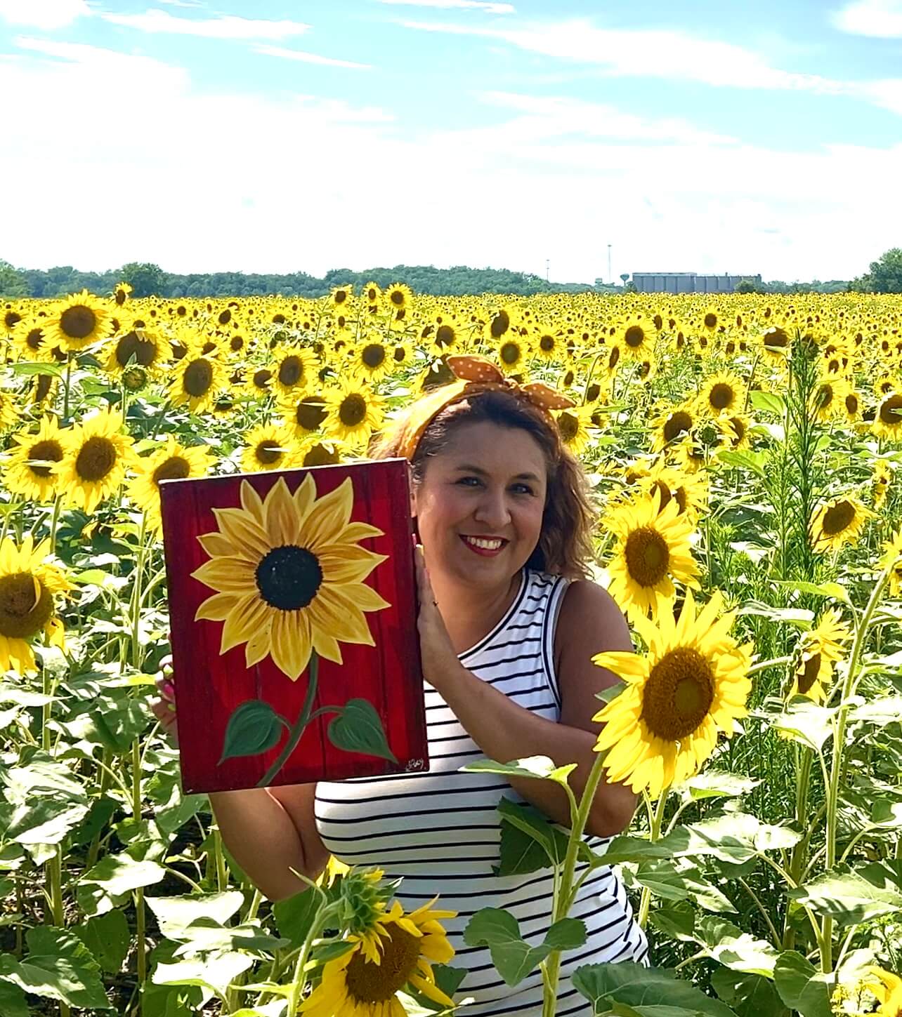 Image of Lucy Schmidt, owner of Lucy's Painting Gallery, holding a painting of a sunflower while standing in a field of sunflowers