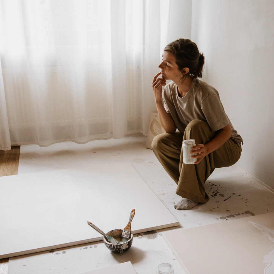 Christina Hohenwarter crouching in her light-filled studio beside blank canvases and a bowl of brushes, looking toward the window.